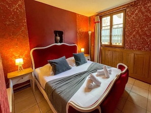 Habitación de hotel con pared de acento roja, cama con ropa blanca, dos almohadas, mesita de noche con lámpara y ventana con cortinas.