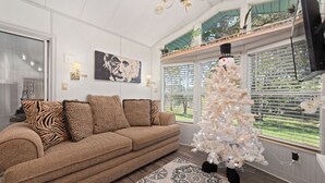 A living room with a brown sofa, leopard print pillows, a white Christmas tree, and a large window with blinds.