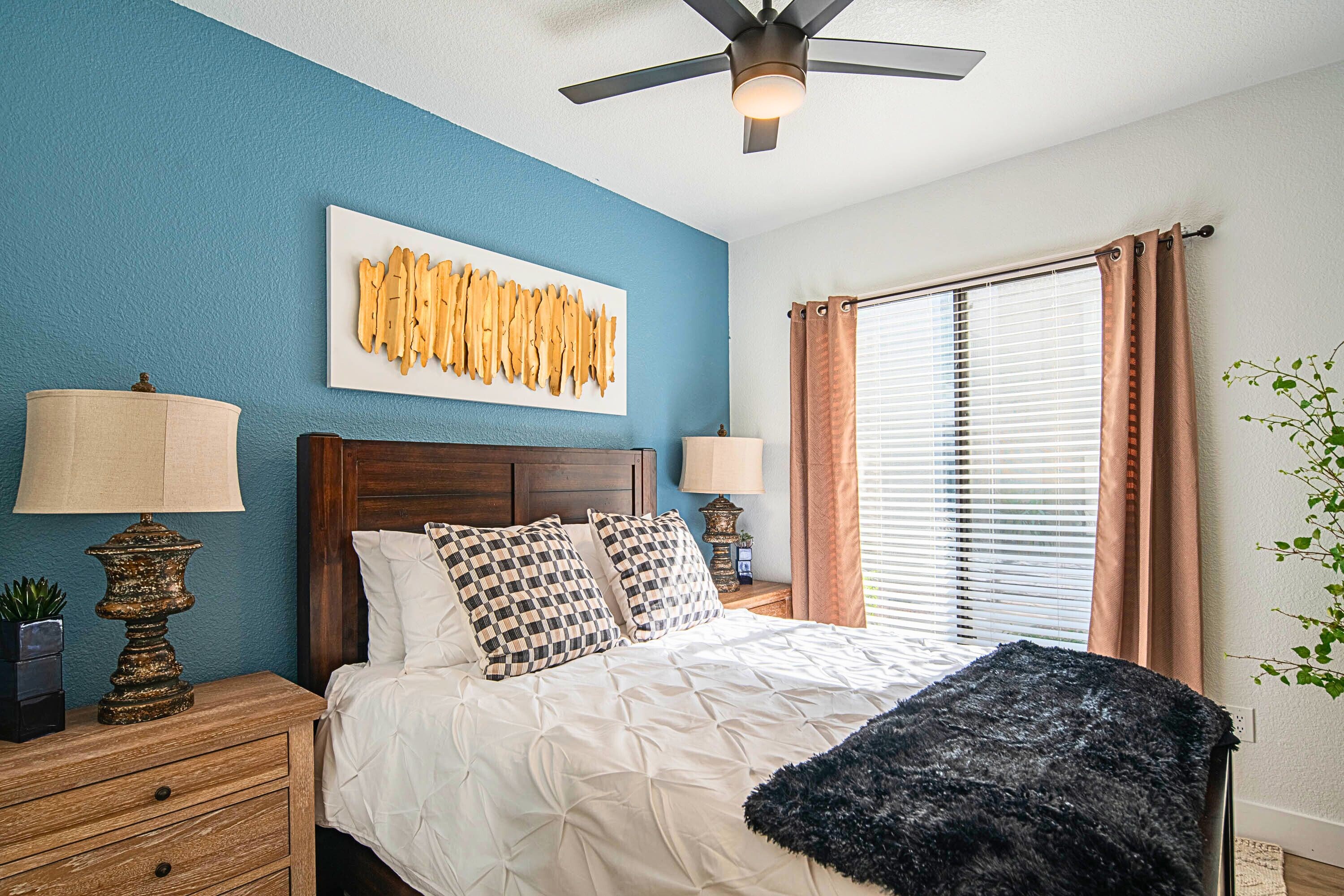 A bedroom with a bed, two nightstands, a ceiling fan, a window with blinds, and a wall art piece.