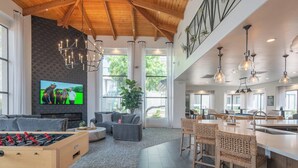 A spacious living area with a high ceiling, a large TV, a foosball table, and a kitchen area with bar stools.