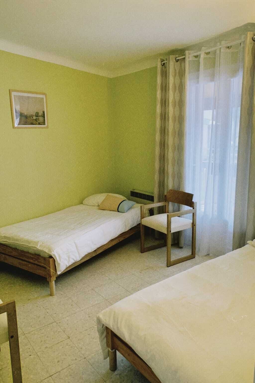 A room with two single beds, a chair, a radiator, and a window with curtains.