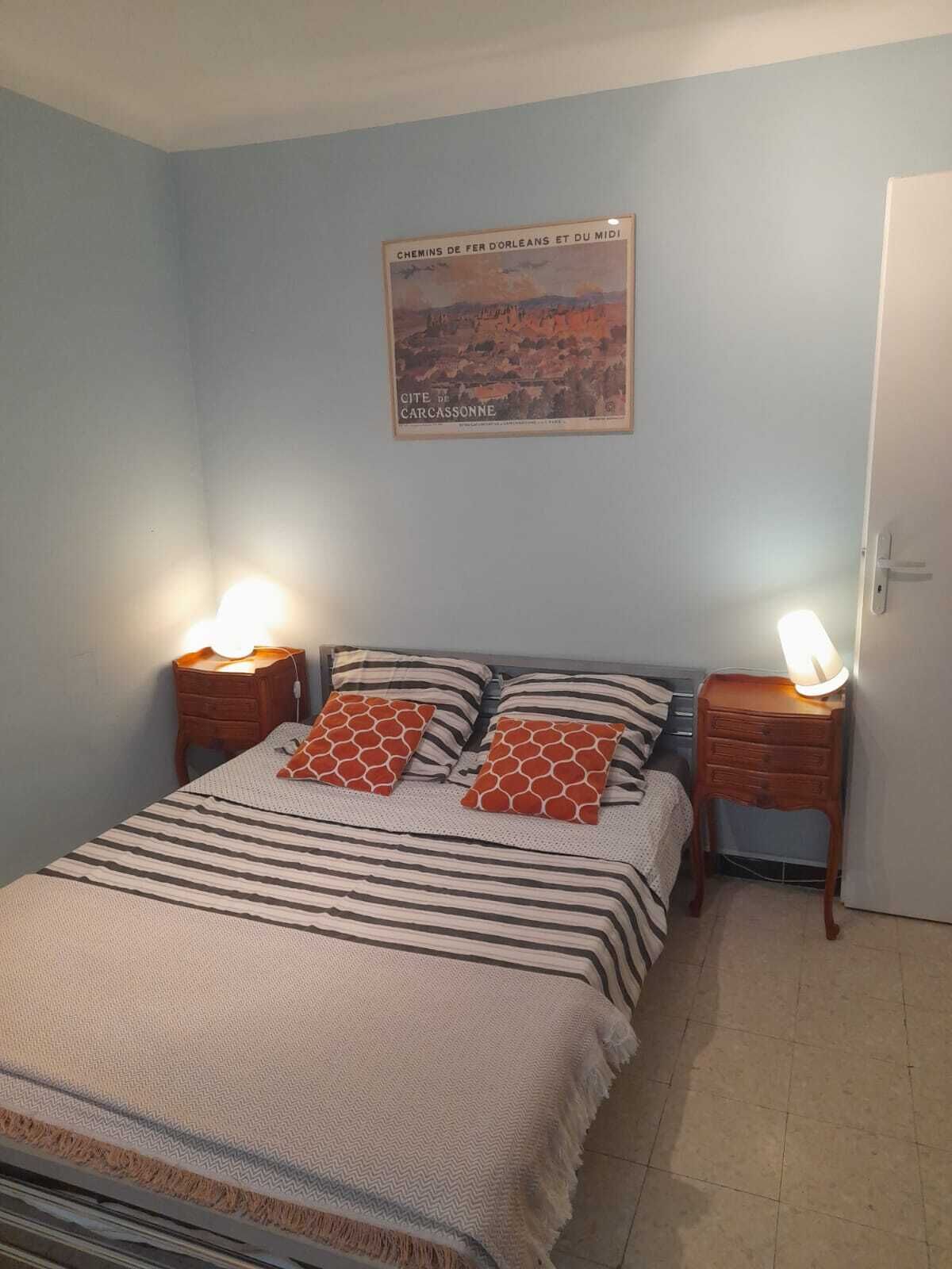 A bedroom with a bed, two bedside tables, a nightstand, a door, and a wall-mounted poster.