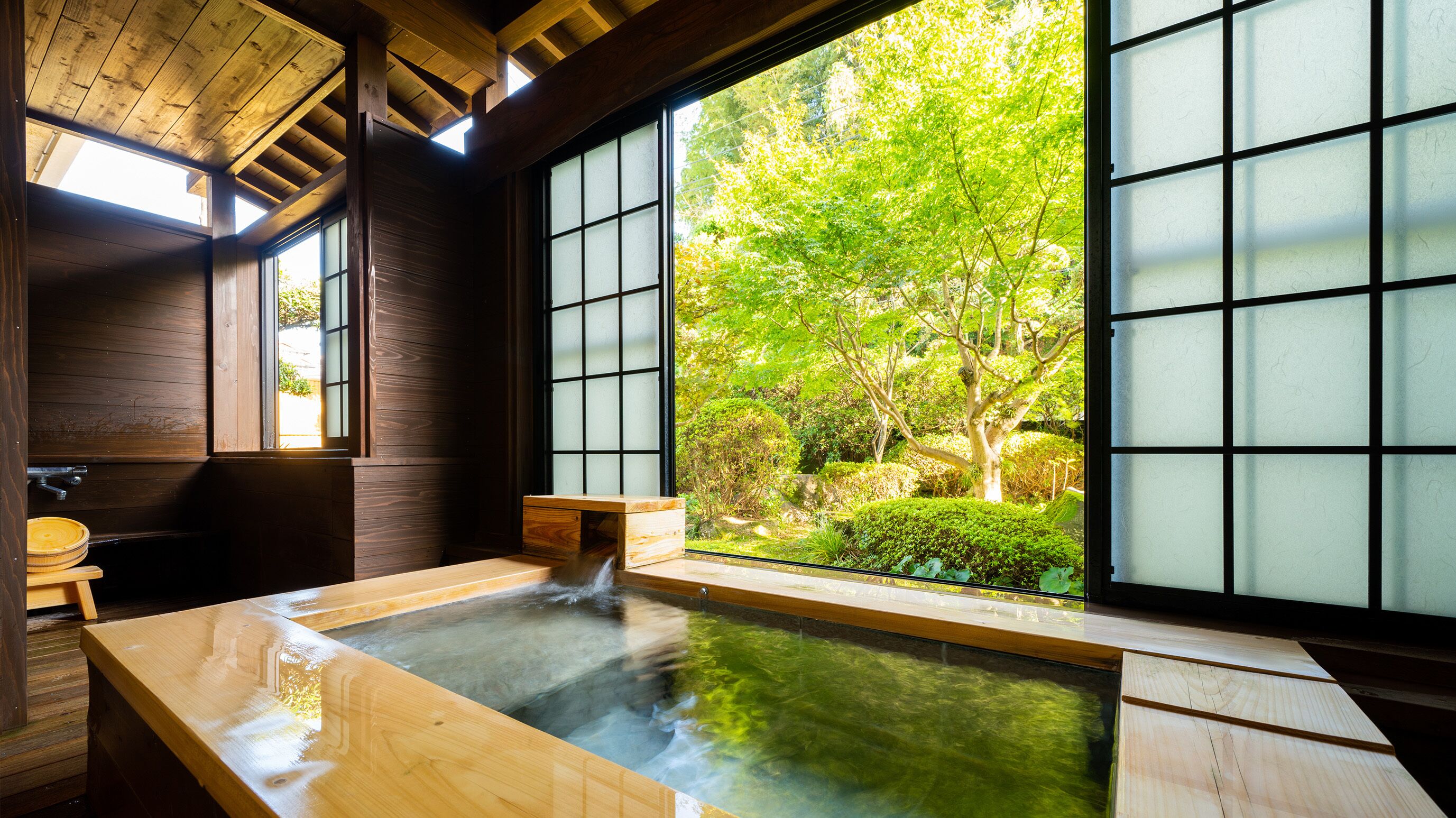 Un onsen japonès tradicional amb interiors de fusta, una banyera de fusta i vista a un jardí exuberant a través de grans portes corredisses.