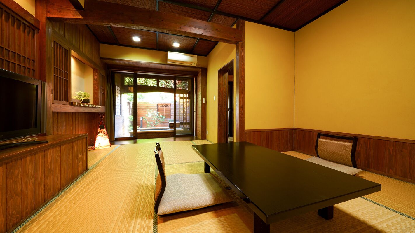 A tatami mat room with a low table and chairs, wooden paneling, and a sliding door leading to an outdoor area.
