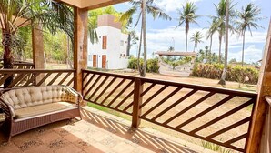 Een overdekt terras met een comfortabele bank en houten balustrade, met uitzicht op een wit gebouw en palmbomen.