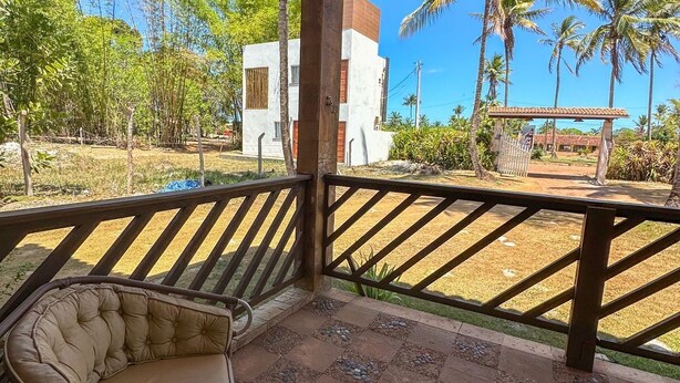 Een overdekt terras met een rieten stoel en een tafel, omringd door een houten hekwerk en uitzicht op een wit gebouw en palmbomen.