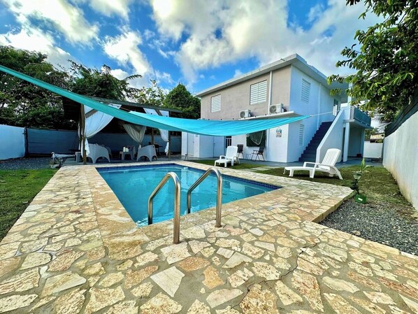 A swimming pool with stone tiles, a covered seating area, and a two-story building with air conditioning units.