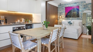 Moderni keittiö, jossa on valkoiset kaapit, puinen ruokapöytä ja tuolit. Siellä on pieni ruokailutila, jossa on valkoinen sohva ja seinällä taulu.