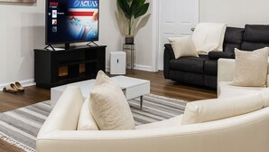 Ett modernt vardagsrum med en vit lädersoffa, en svart lädersoffa, en platt-TV, en krukväxt och en vit dörr.