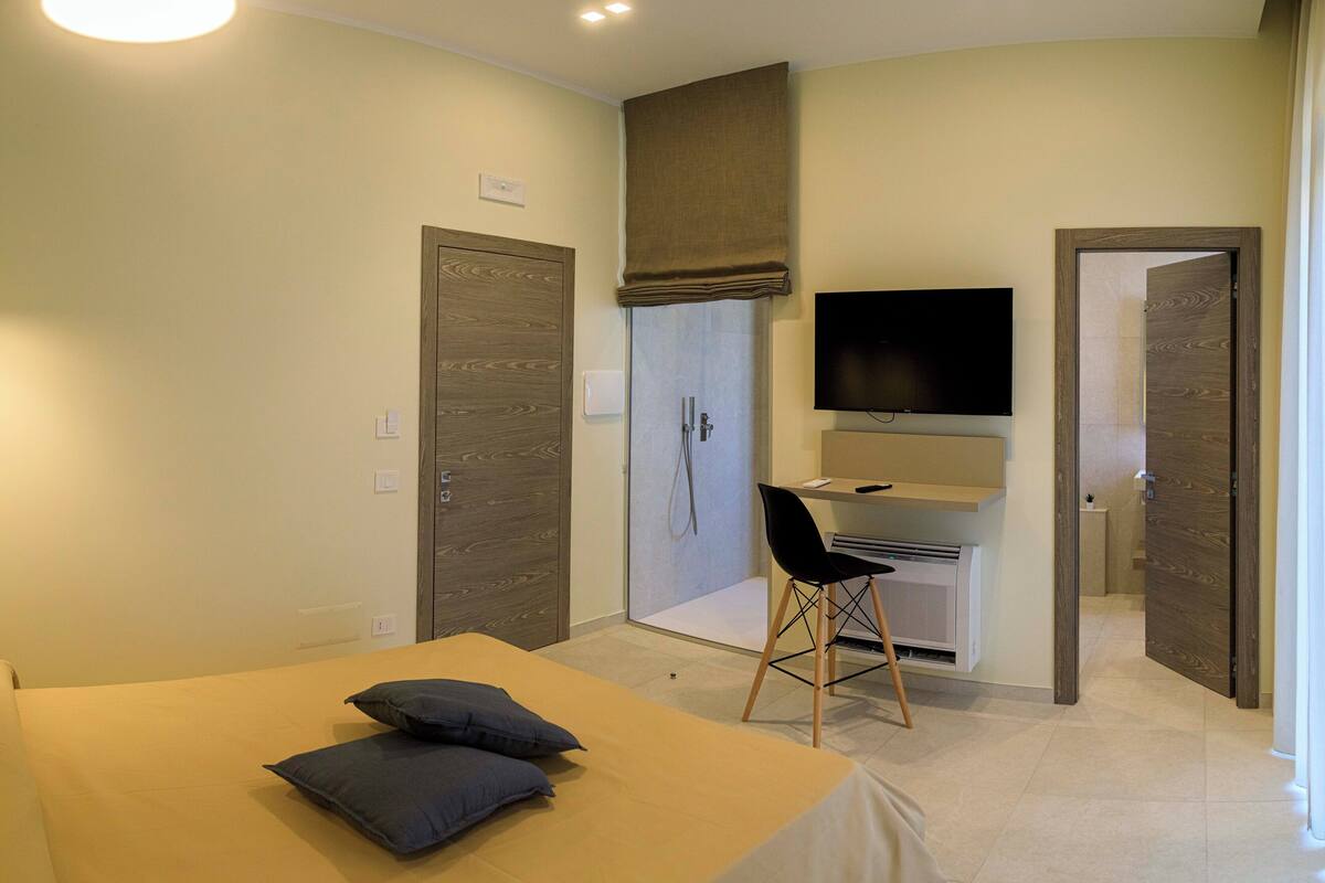 A bedroom with a bed, a desk, a chair, a TV, and a shower area.