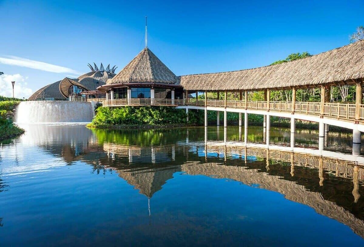 A resort with a thatched-roof pavilion, a water feature, and a bridge over a calm body of water.