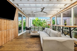 Eine überdachte Terrasse im Freien mit Holzdeck, einer Couch, einem Tisch, Stühlen und Blumentöpfen.