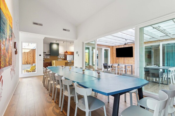 A spacious dining area with a long table and multiple chairs, a kitchen in the background, and a large window providing natural light. - The Cocodrie at CastleDay- Private Pool, Spa (New Orleans)