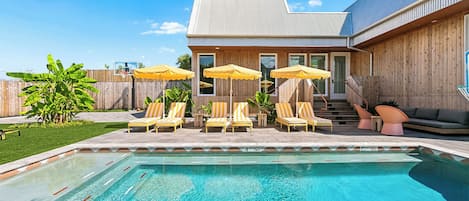 A swimming pool with lounge chairs and umbrellas, a wooden fence, and a building with a sloped roof.