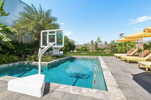 A swimming pool with a basketball hoop, lounge chairs, and umbrellas.