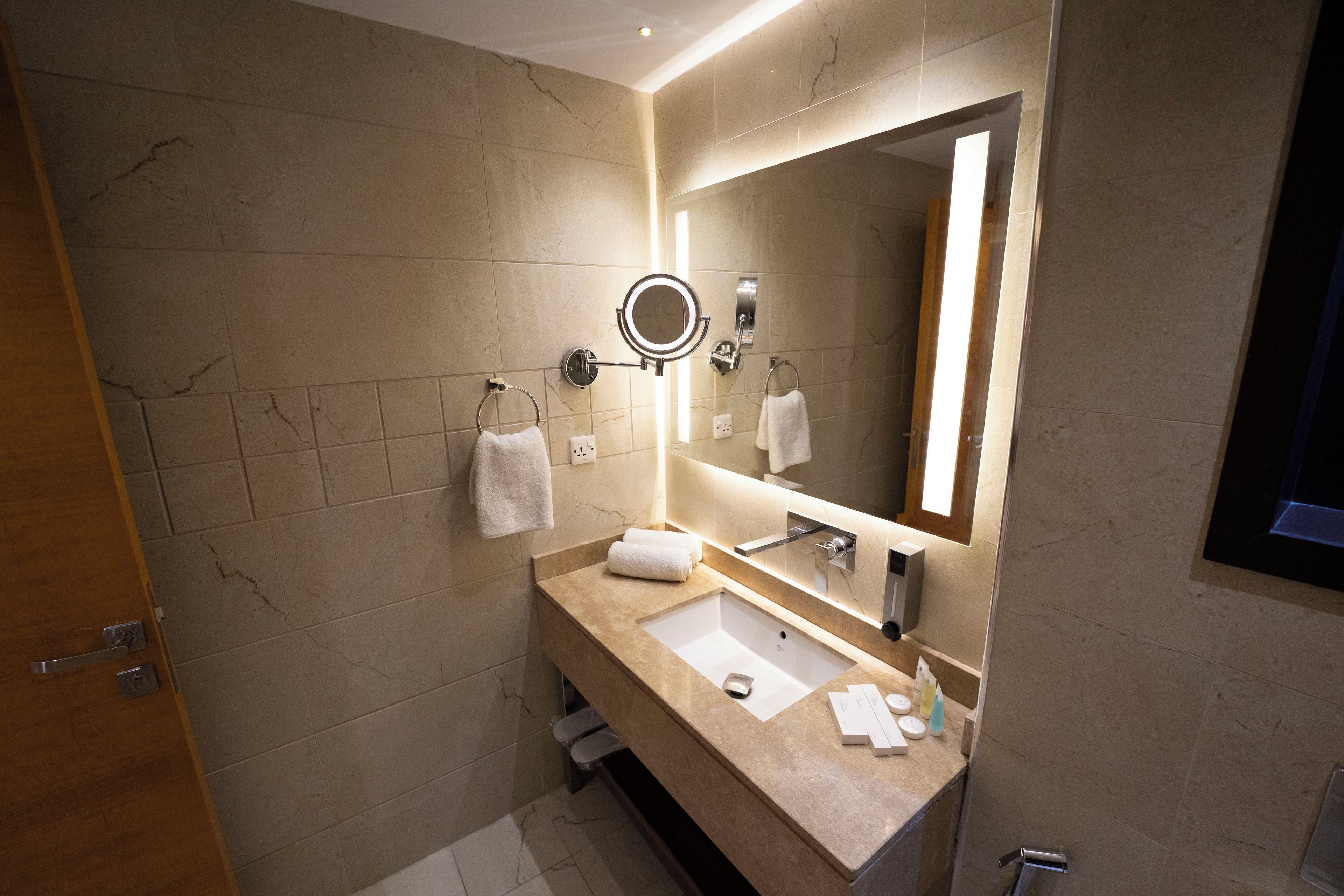 A modern bathroom with a marble-tiled wall, a large mirror with integrated lighting, a sink with a vanity, and a towel rack.