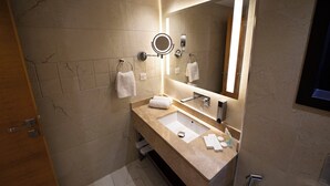 A modern bathroom with a marble-tiled wall, a large mirror with integrated lighting, a sink with a vanity, and a towel rack. - Gloria Boutique Hotel (Amman)