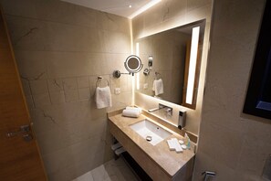 A modern bathroom with a marble-tiled wall, a large mirror with integrated lighting, a sink with a vanity, and a towel rack. - Gloria Boutique Hotel (Amman)