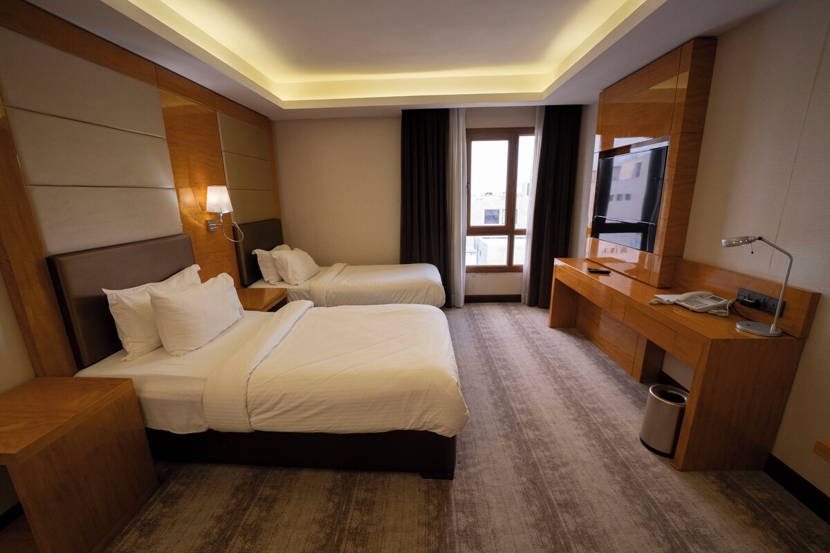 A hotel room with two beds, a desk, a TV, and a window with curtains.