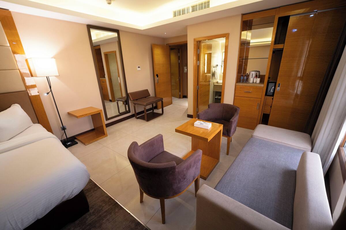 A hotel room with two beds, a sofa, a small table, and a chair. There is a mirror, a lamp, and a wardrobe.