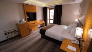 Una habitación de hotel con cama, mesita de noche, televisor, escritorio de madera y ventana con cortinas.