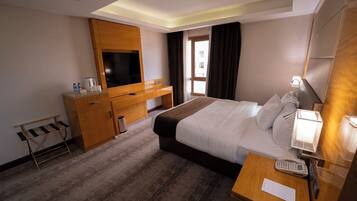 A hotel room with a bed, bedside table, TV, wooden desk, and a window with curtains.