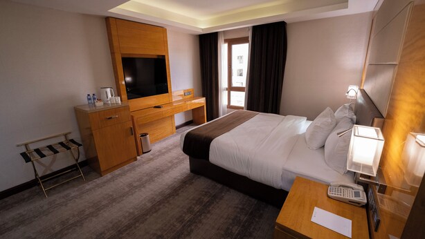 A hotel room with a bed, bedside table, TV, wooden desk, and a window with curtains. - Gloria Boutique Hotel (Amman)
