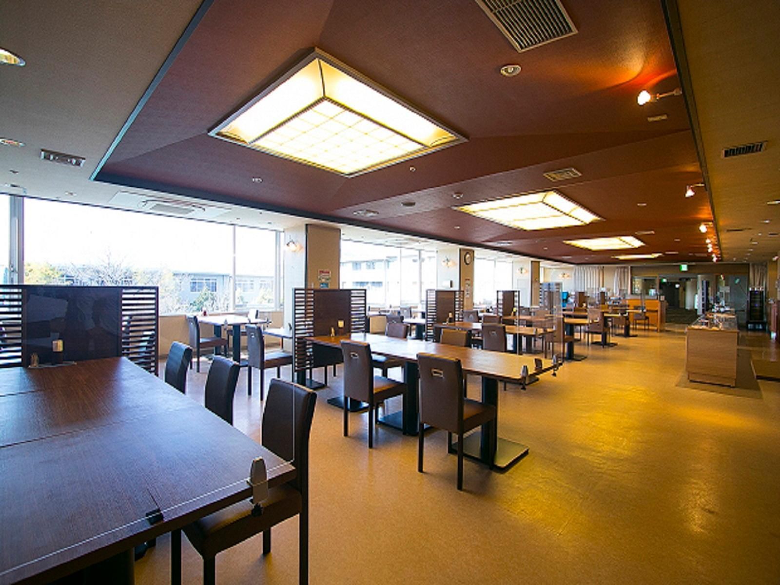 A spacious indoor area with multiple tables and chairs, large windows allowing natural light, and a modern ceiling design.