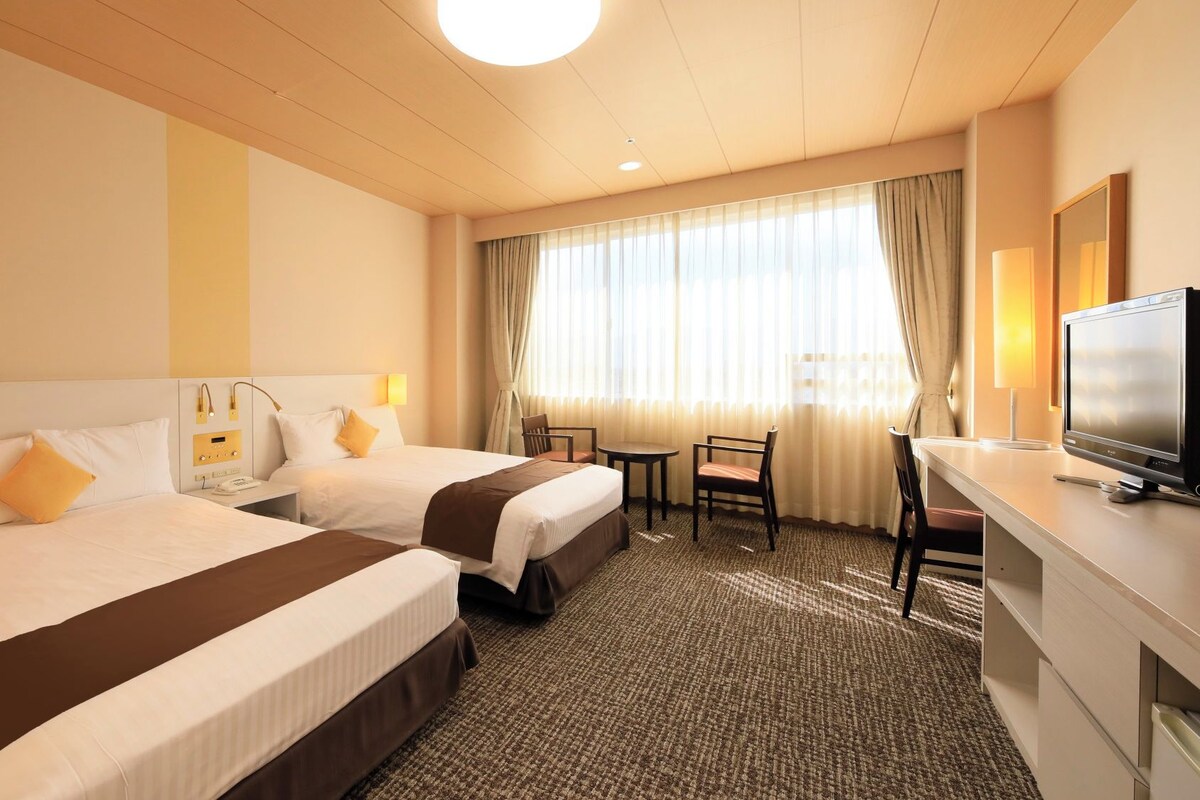 A hotel room with two beds, a desk, a television, and a window with curtains.