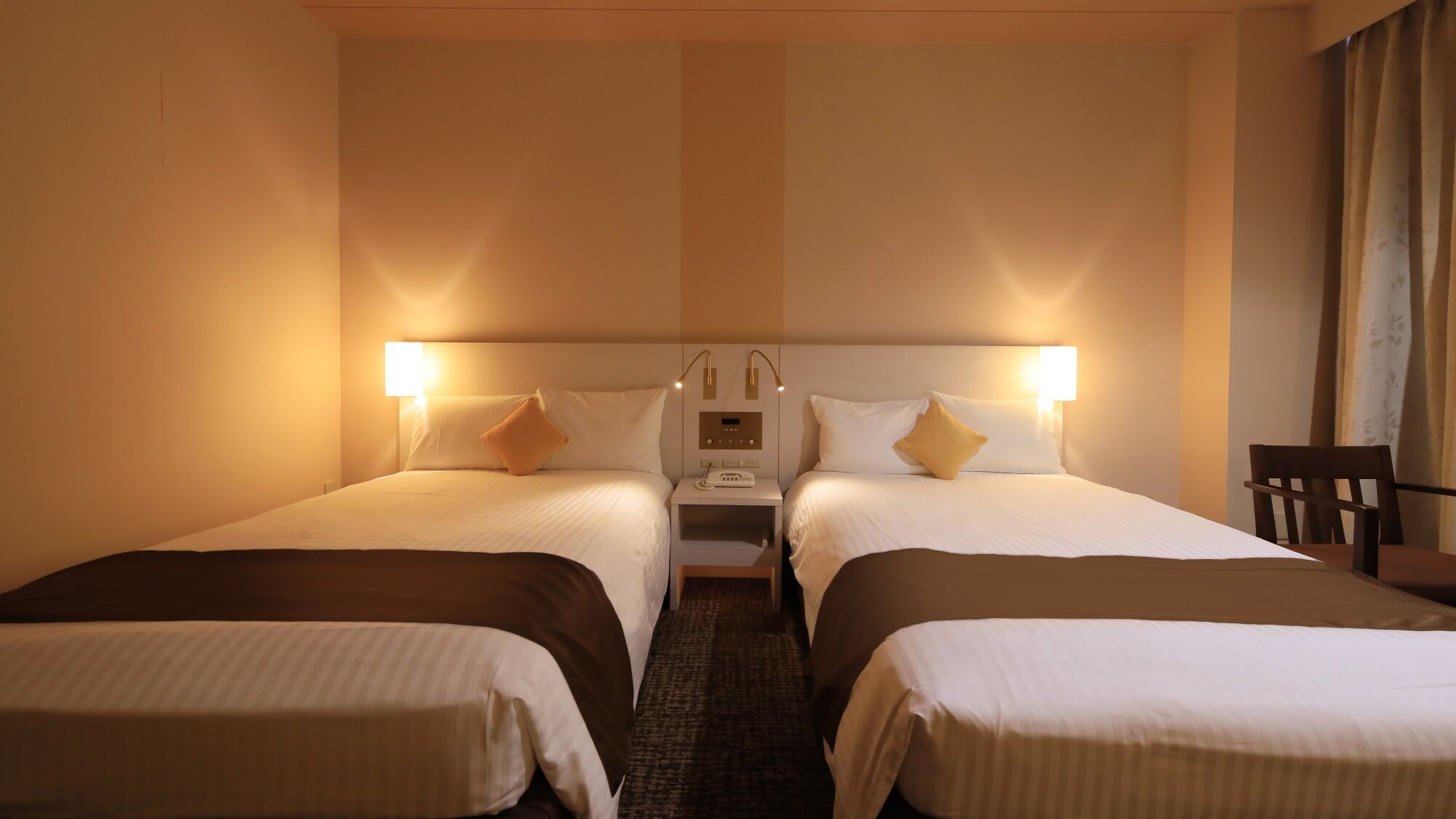 Ett hotellrum med två separata sängar, ett nattduksbord med en lampa, en kontrollpanel och en stol.