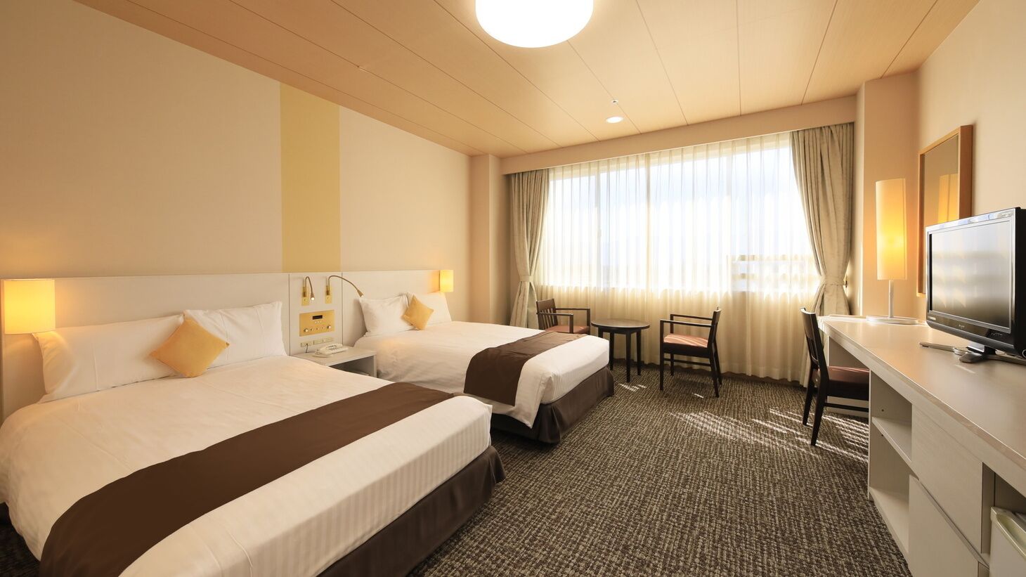 Ett hotellrum med två sängar, ett skrivbord med en tv, en stol, ett litet bord och ett fönster med gardiner.