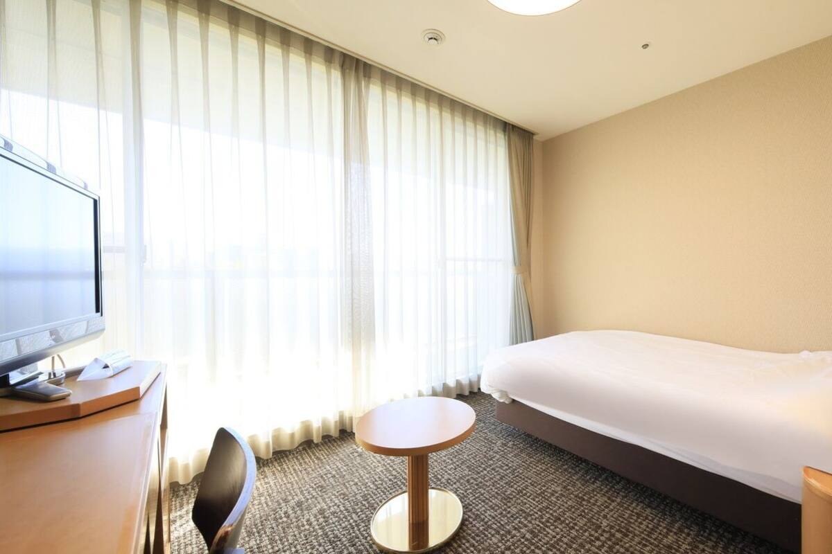 Habitación de hotel con cama, escritorio, computadora, silla, lámpara y ventana con cortinas.