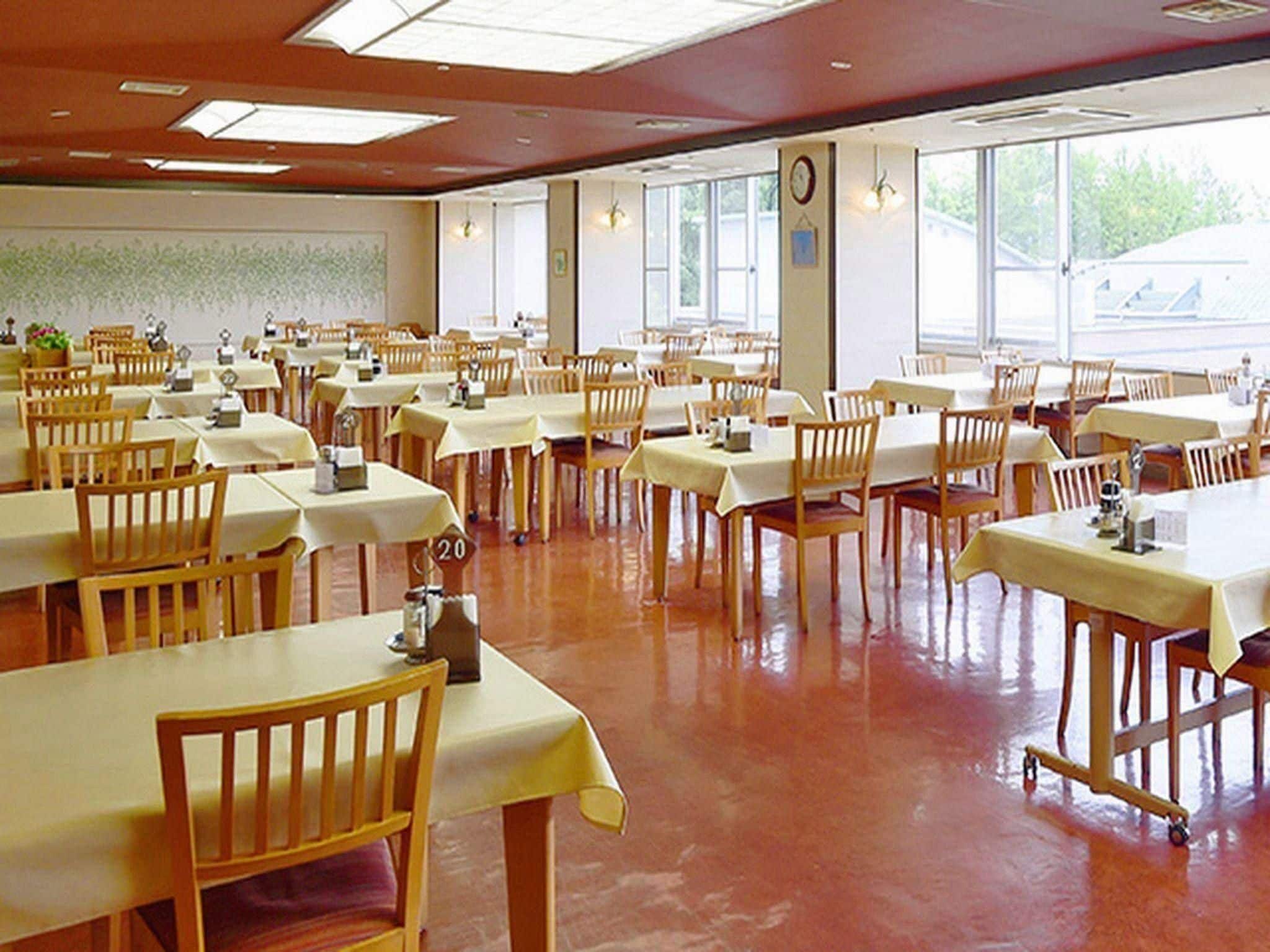 A spacious dining hall with multiple tables and chairs, large windows, and a clock on the wall.