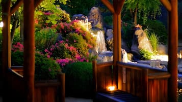 A wooden gazebo with a bench, surrounded by lush greenery and a small waterfall.
