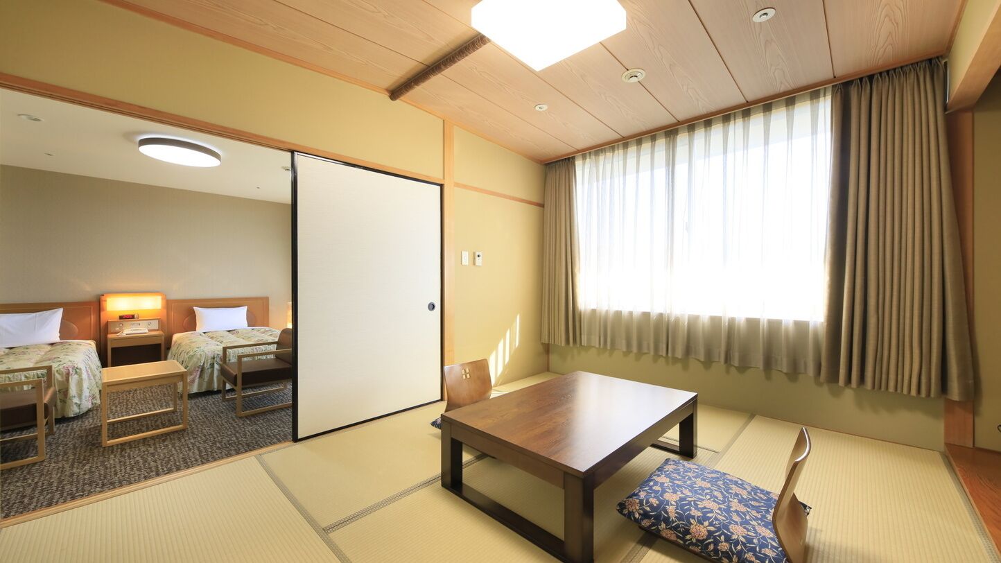 Una habitación japonesa tradicional con suelo de tatami, una mesa de madera, dos camas con almohadas, una puerta corredera y un ventanal con cortinas.