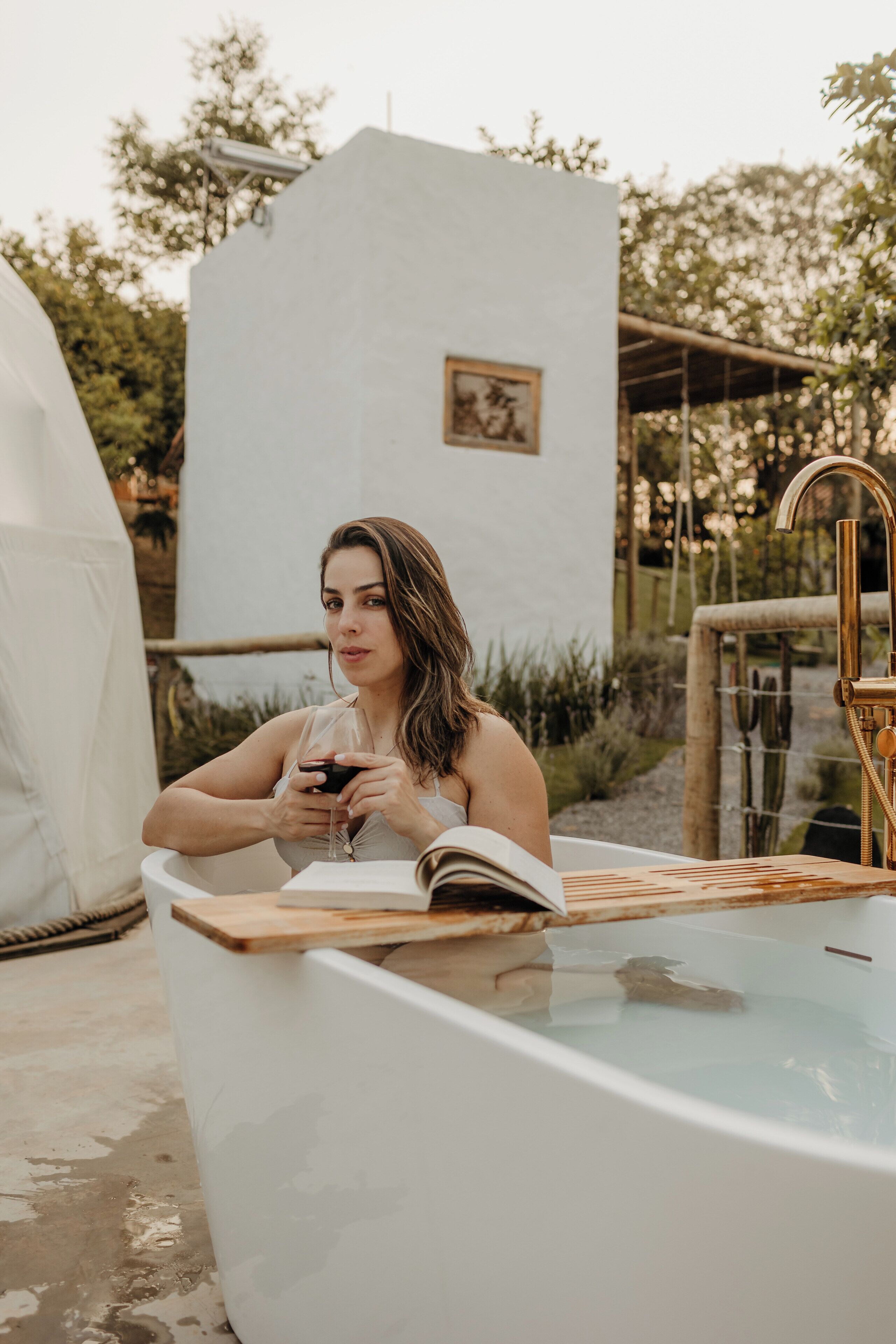 A person relaxing in a bathtub with a book and a glass of wine, surrounded by a garden and a white building.