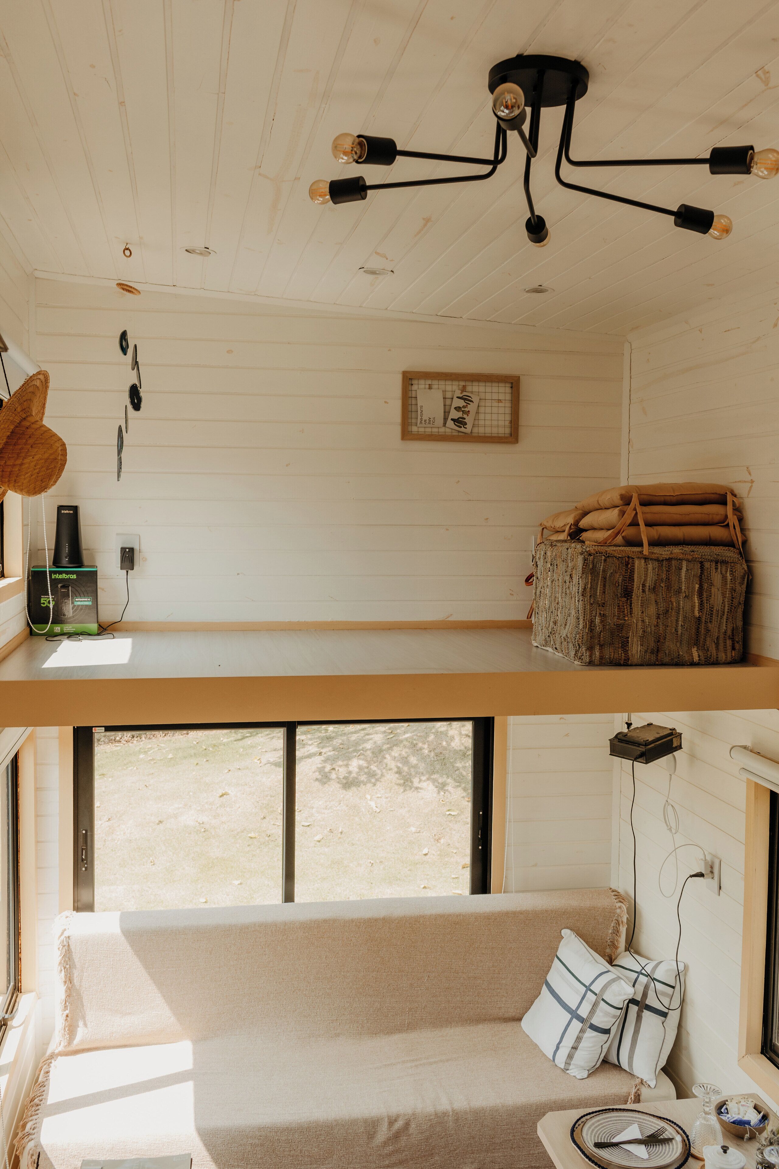 A cabin interior with a loft bed, a sofa, a hanging light fixture, and a window overlooking a natural landscape.
