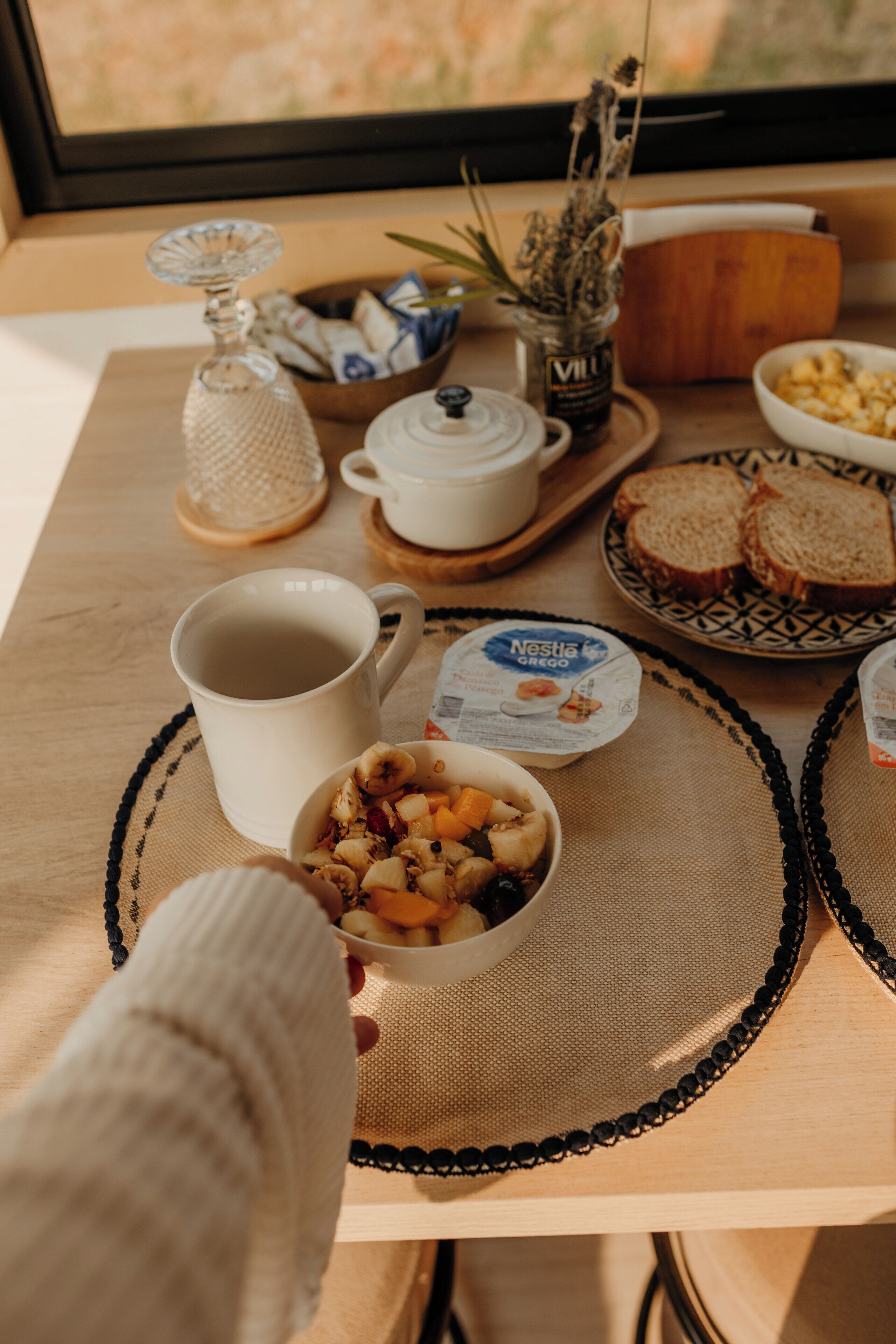 A breakfast table with a cup, bowl of cereal, bread, and a jar of preserves.
