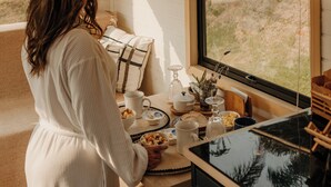 A woman in a white dress standing by a kitchen counter with a view of a grassy landscape through a large window. - Dila's Lodge (Sao Roque)