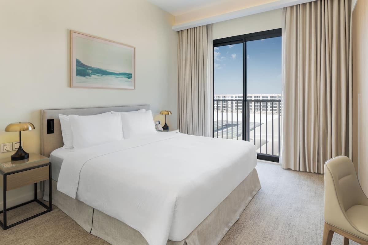 A hotel room with a large bed, a nightstand with a lamp, a chair, a painting on the wall, and a view of the city through the window.