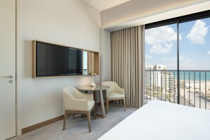 A hotel room with a bed, a TV, a desk with chairs, and a view of a beach and buildings. - Vida Beach Resort Marassi Al Bahrain (Manama)