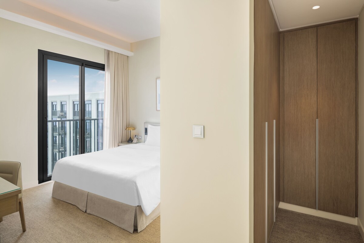 A hotel room with a large bed, a desk, a chair, a window with a view of buildings, and a wardrobe.