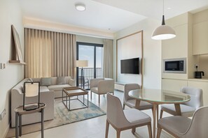 A modern hotel room with a sofa, chairs, a glass dining table, a TV, and a balcony with curtains. - Vida Beach Resort Marassi Al Bahrain (Manama)