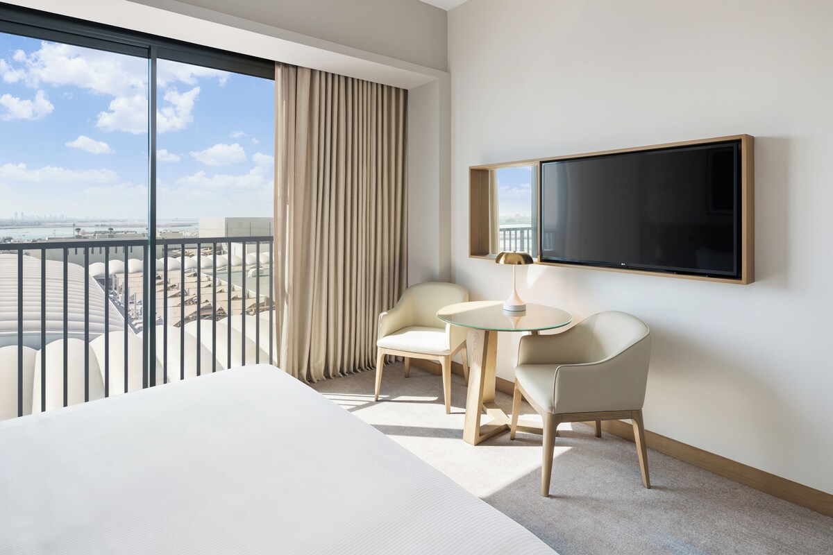 A hotel room with a large window, a bed, a TV, and a small table with chairs.