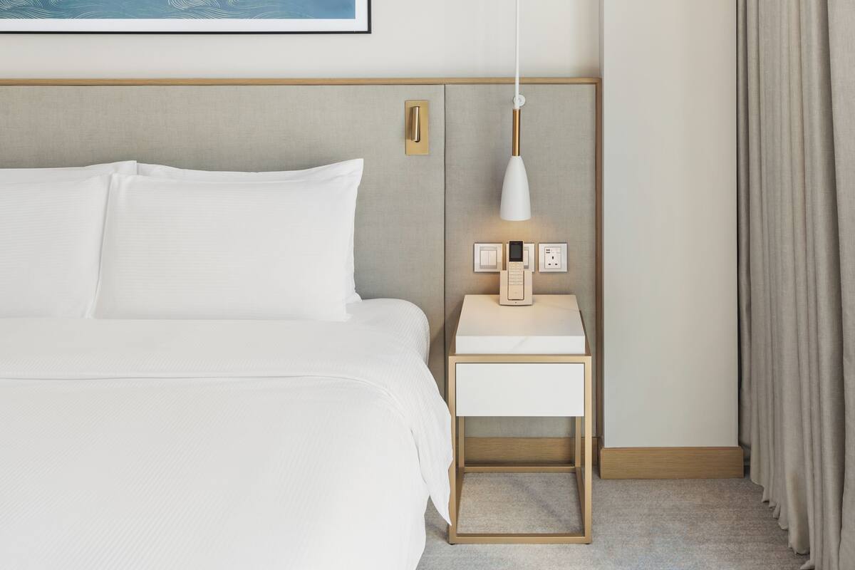 A neatly made bed with white linens, a bedside table with a lamp, and a wall-mounted light fixture.