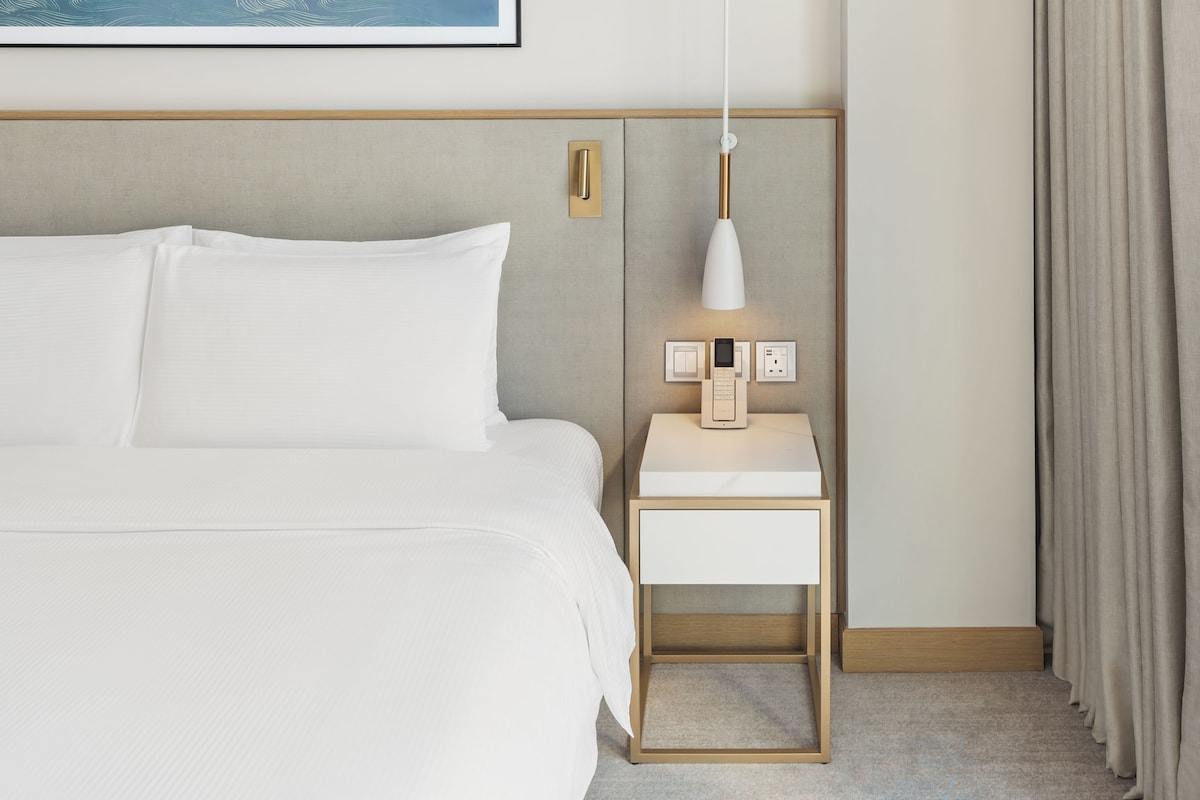 A neatly made bed with white linens, a bedside table with a lamp, and a wall-mounted light fixture.
