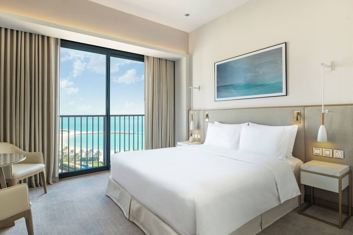 A hotel room with a large bed, a bedside table, a chair, a desk, and a view of the ocean.