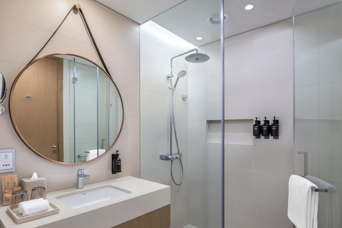 A modern bathroom with a round mirror, a sink, a shower, and toiletries on the wall.