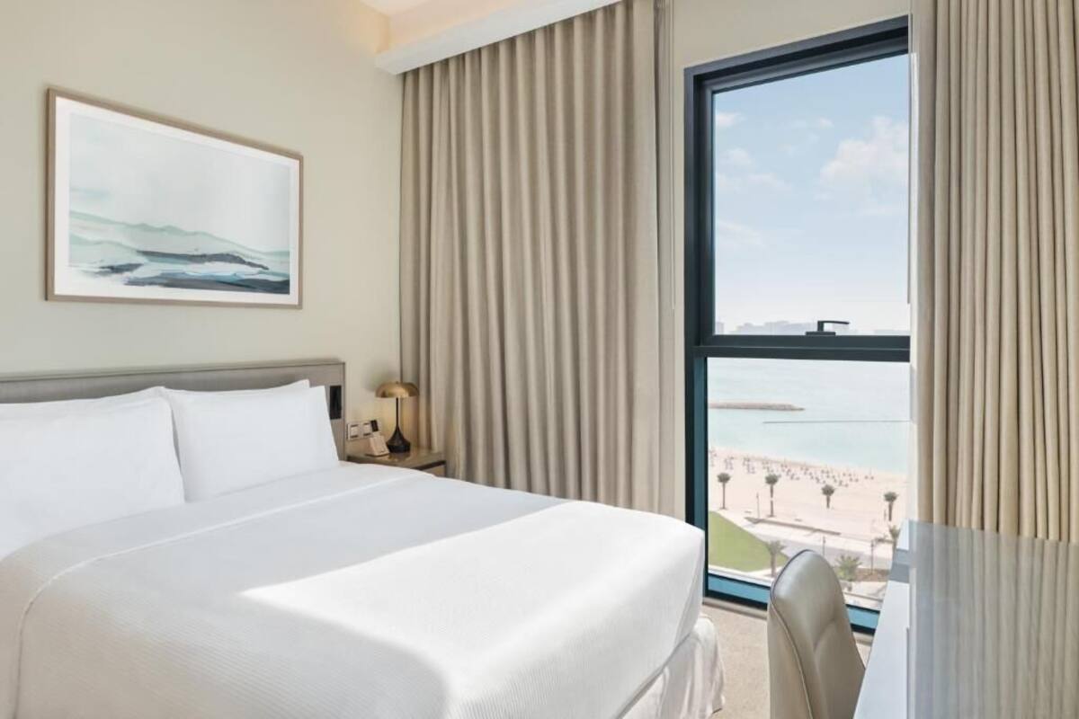A hotel room with a bed, a nightstand with a lamp, a large window with a view of a beach and water, and a framed picture on the wall.