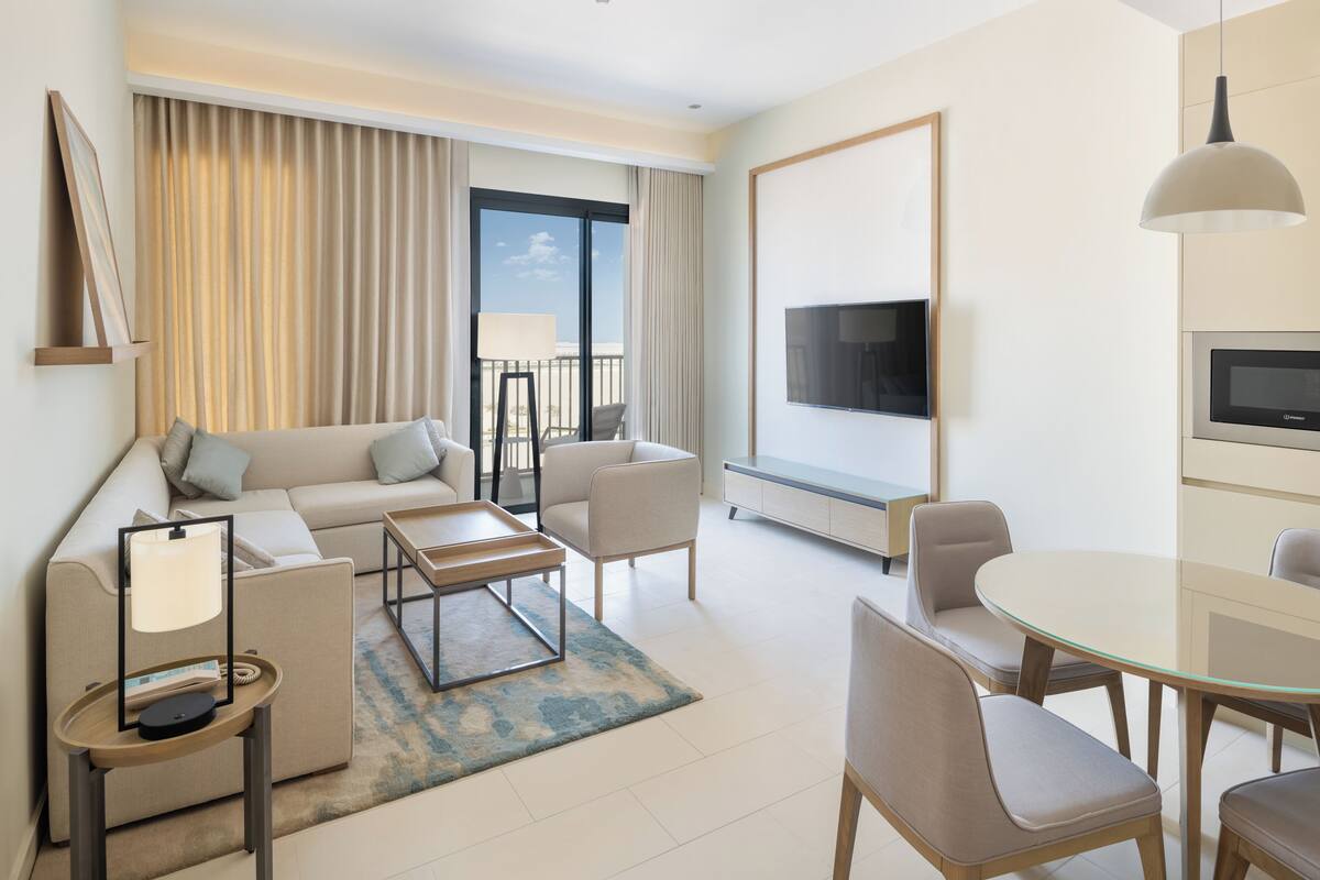 A modern hotel room with a sofa, a coffee table, a dining table with chairs, a TV, and a balcony with curtains.